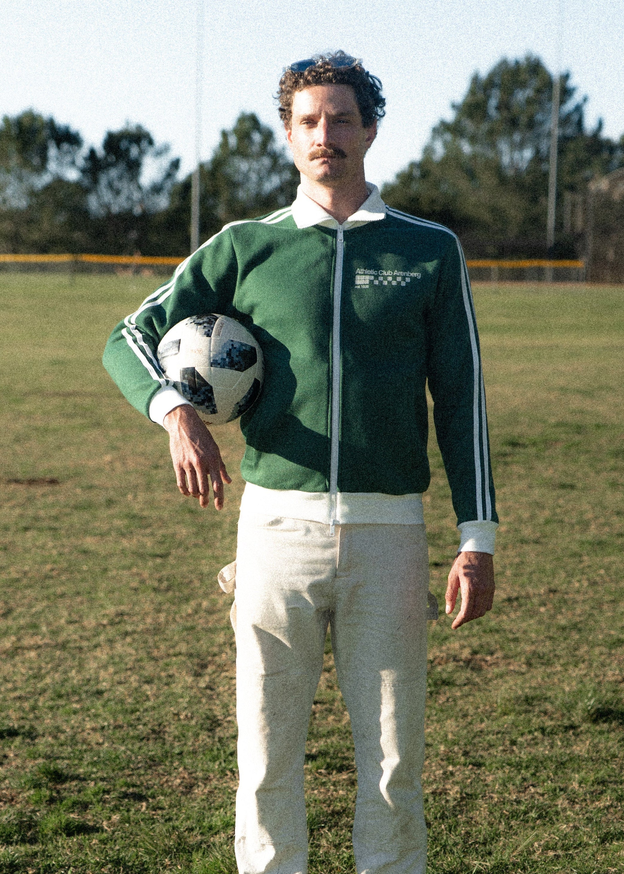 Man in a green jacket holding a soccer ball on a grassy field