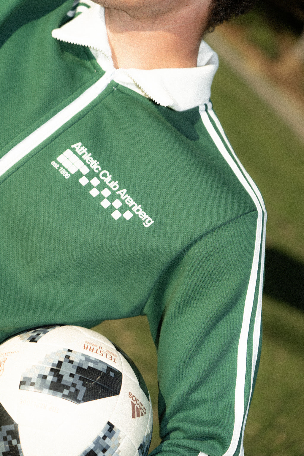 Person wearing a green tracksuit with a logo, holding a soccer ball on a grassy field.