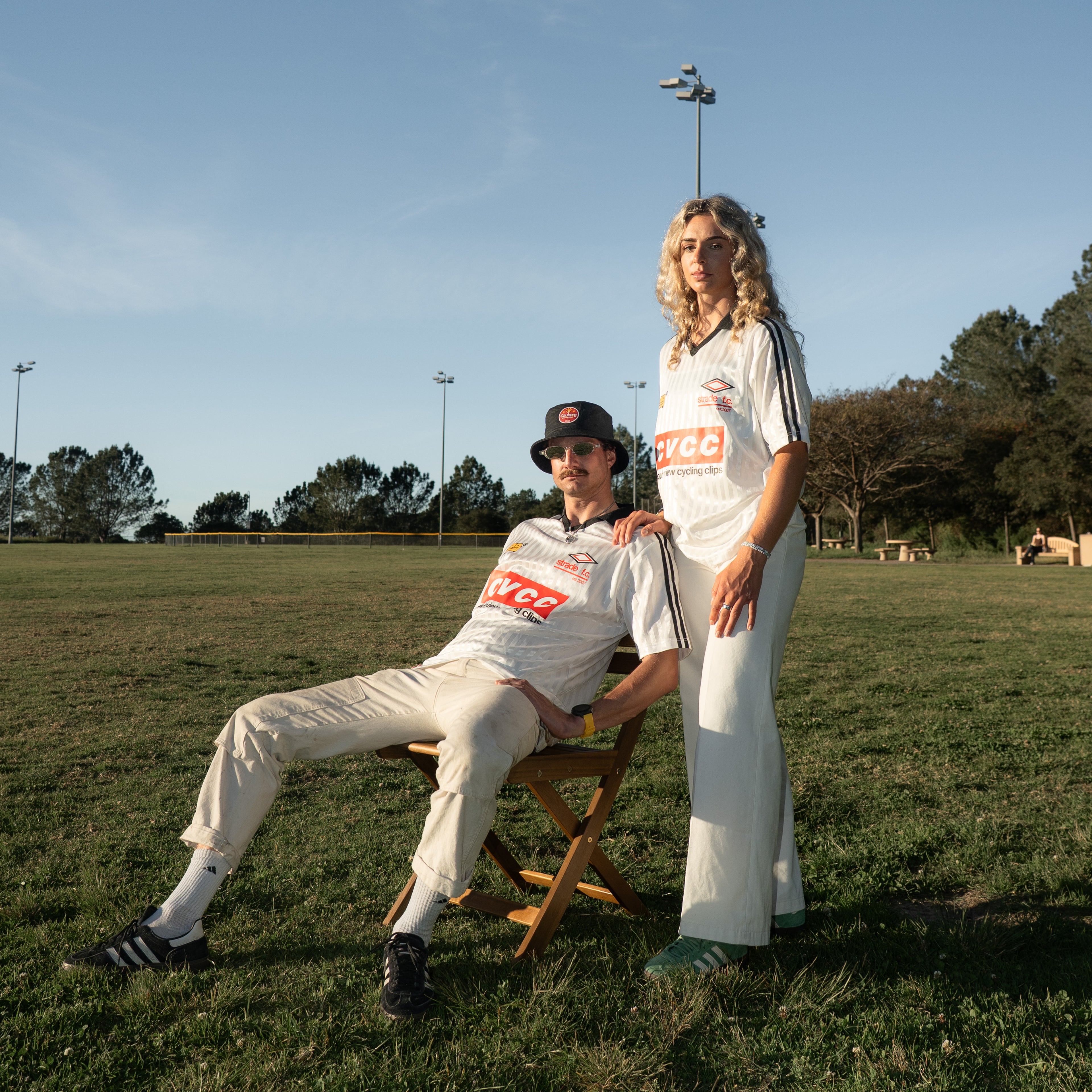 Two people in sports attire sitting on a chair in a grassy field with trees and clear sky.